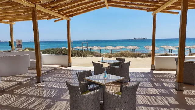 A reed pergola with grey wicker armchairs, facing a sandy beach, straw parasols and a turquoise sea