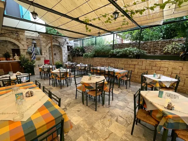 A covered courtyard with orange checked tablecloths, green metal chairs, a stone wall and a bust