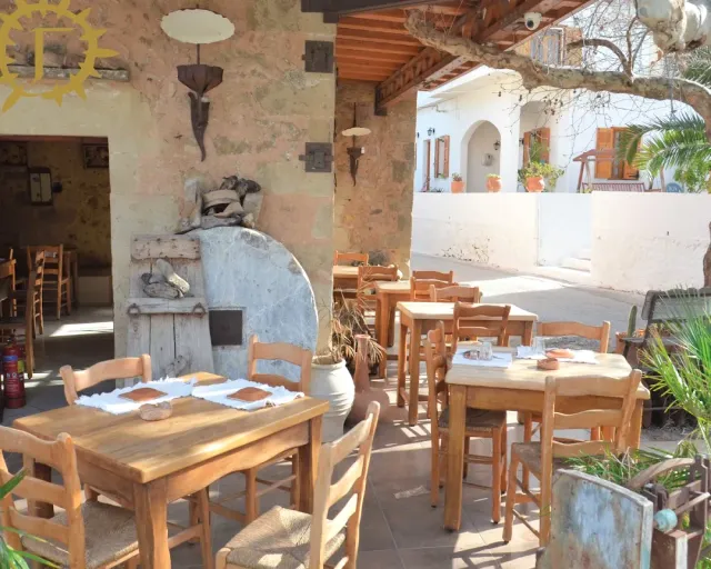 A covered terrace with a stone wall and an old oven, wooden tables and straw chairs in the sun