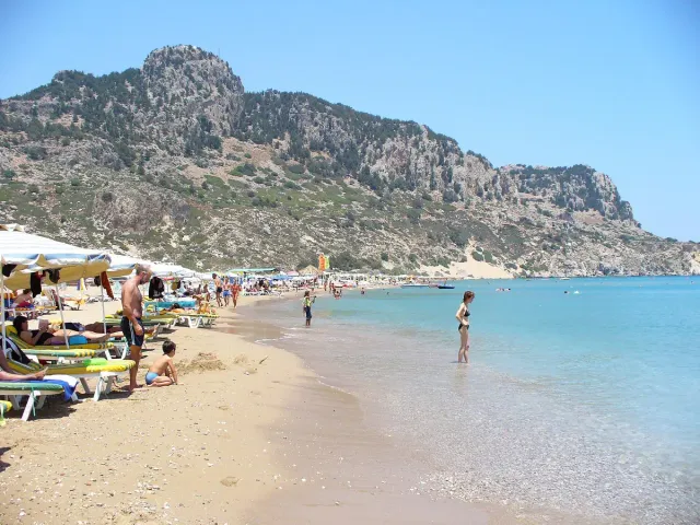 A sandy beach with sunbeds and bathers, backed by a high rocky mountain
