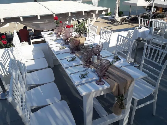 A long table set with pink glasses, red roses and white chairs facing the sea