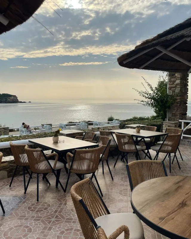 Tables and wicker chairs under thatched shades, facing a calm sea and a cloudy sky