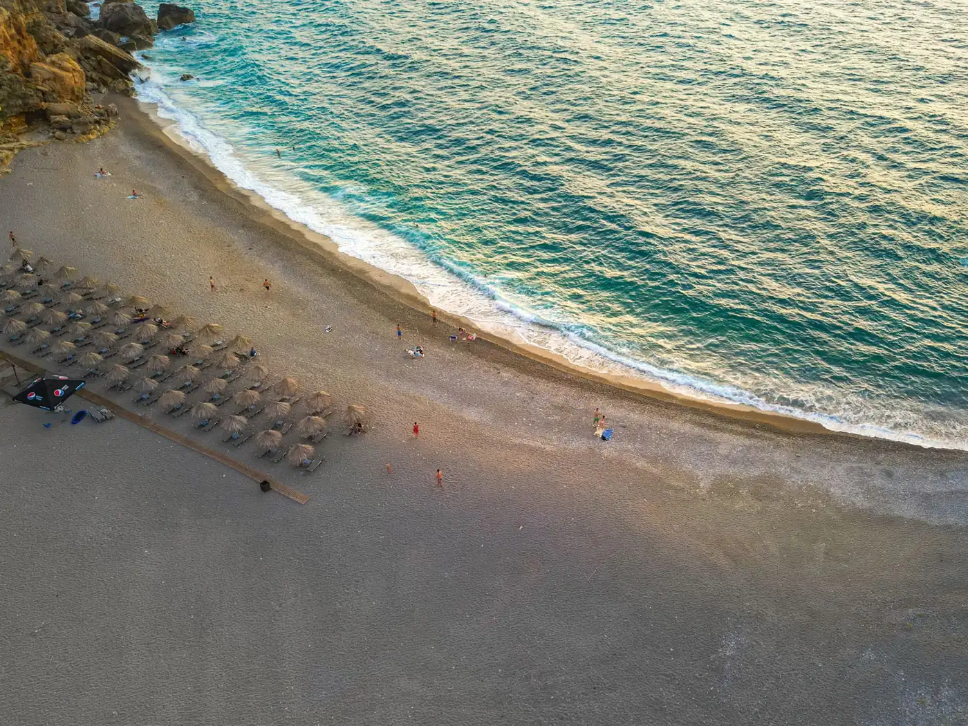 An aerial view of a beach on the coast of Crete with turquoise shallows and rows of parasols at sunset