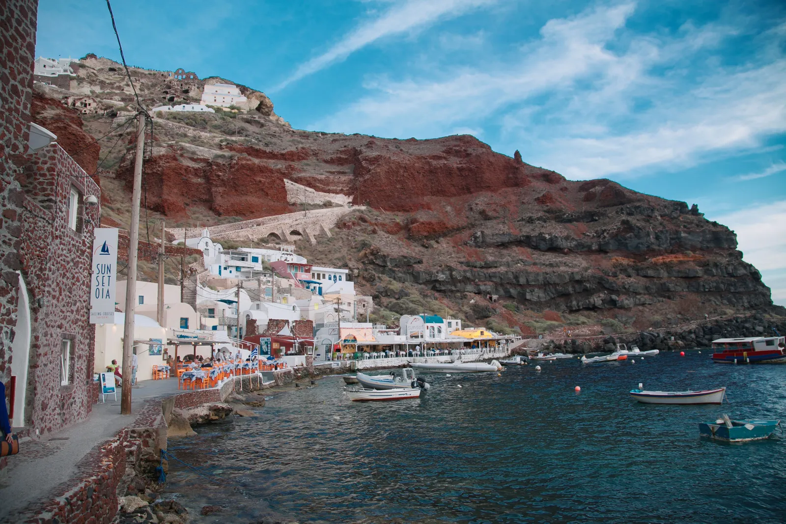 Whitewashed buildings and moored boats at the water's edge below a red cliff in a small bay