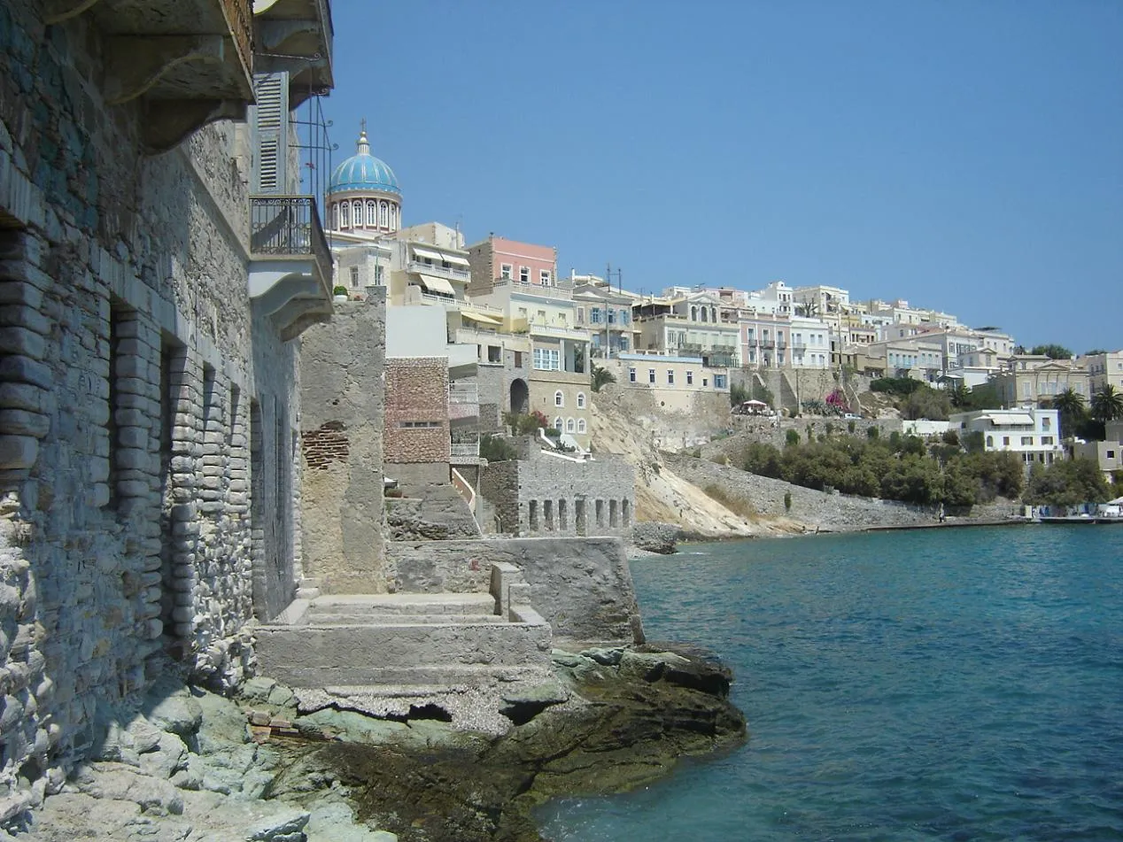 Weathered stone ruins lead the eye toward pastel neoclassical houses and a blue domed church along the Ermoupoli waterfront, Syros, Greece